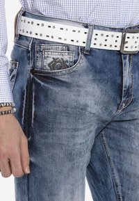 Blauwe spijkerbroek met een vervaagde afwerking, met een zwart logo op de voorkzak. Gecombineerd met een witte gesp-stoere riem en een geruit shirt.