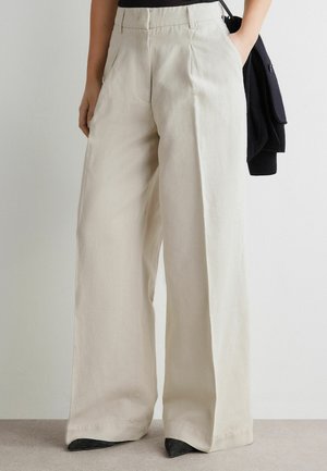 Hochtaillierte, weit geschnittene cremefarbene Hose mit vorderen Falten, kombiniert mit schwarzen Schuhen und einem schwarzen Pullover, der über dem Arm getragen wird.