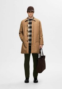 Impermeabile khaki con chiusura a bottoni, indossato sopra una camicia a scacchi. Abbinato a pantaloni verde scuro e con una borsa tote marrone scuro. Cappello marrone.