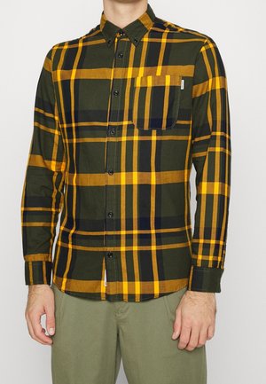 Mann trägt ein geknöpftes, langärmliges Hemd mit einem Karo-Muster in Senfgelb, Schwarz und Dunkelgrün sowie olivgrüne Hose.