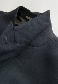 Tumesinise nahkjakk, millel on tekstureeritud viimistlus, seisva kraega ja peente õmblusteenustega. Jakk sisaldab kontrastset voodrit.