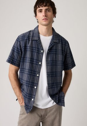 Jeune homme portant une chemise bleu marine à manches courtes avec un motif quadrillé blanc par-dessus un t-shirt blanc, les mains dans les poches d’un pantalon beige.