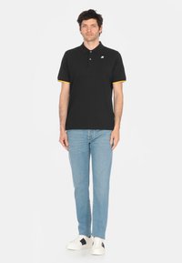 Maglietta polo nera con accenti gialli alle maniche, maniche corte, colletto con bottoni, indossata con jeans azzurri chiari e sneakers bianche.