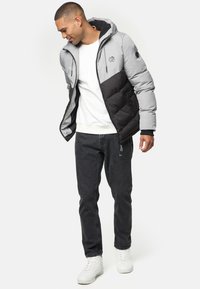 Chaqueta acolchada gris y negra con capucha, cierre frontal con cremallera, puños elásticos y un logo en el pecho, llevada sobre un sudadera blanca y jeans oscuros.