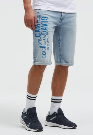 Mann trägt hellblaue Jeansshorts mit aufgerollten Bündchen und blauem Text, weiße Socken mit schwarzen Streifen und dunkelblaue Sneaker.