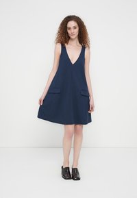 Φόρεμα σε γραμμή Α σε σκούρο μπλε χρώμα, χωρίς μανίκια, με βαθύ V-λαιμόκοψη και δύο μπροστινές τσέπες. Κατασκευασμένο από απαλό, υφασμάτινο ύφασμα με υφή για άνετη εφαρμογή.