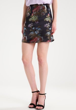 Femme portant une jupe courte noire à motif floral et des sandales noires à talons hauts, debout sur un fond blanc.