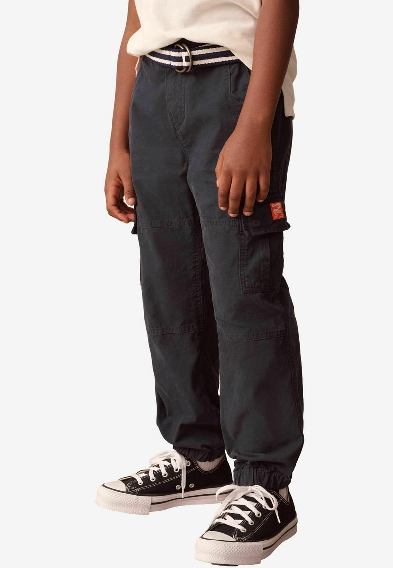 Zwarte cargobroek met een relaxte pasvorm, voorzien van zijzakken en elastische boorden, gecombineerd met zwarte sneakers en een gestreepte riem.