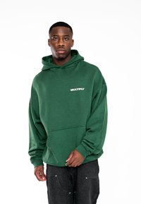 Sudadera verde con un bolsillo frontal, hecha de tejido texturizado, con un logo blanco de "MULTIPLY" en el pecho. Llevada con pantalones negros.