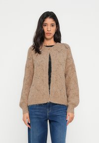 Cardigan beige en tricot avec motif en losange, encolure ronde, fermeture boutonnée et manches volumineuses, porté avec un jean bleu.