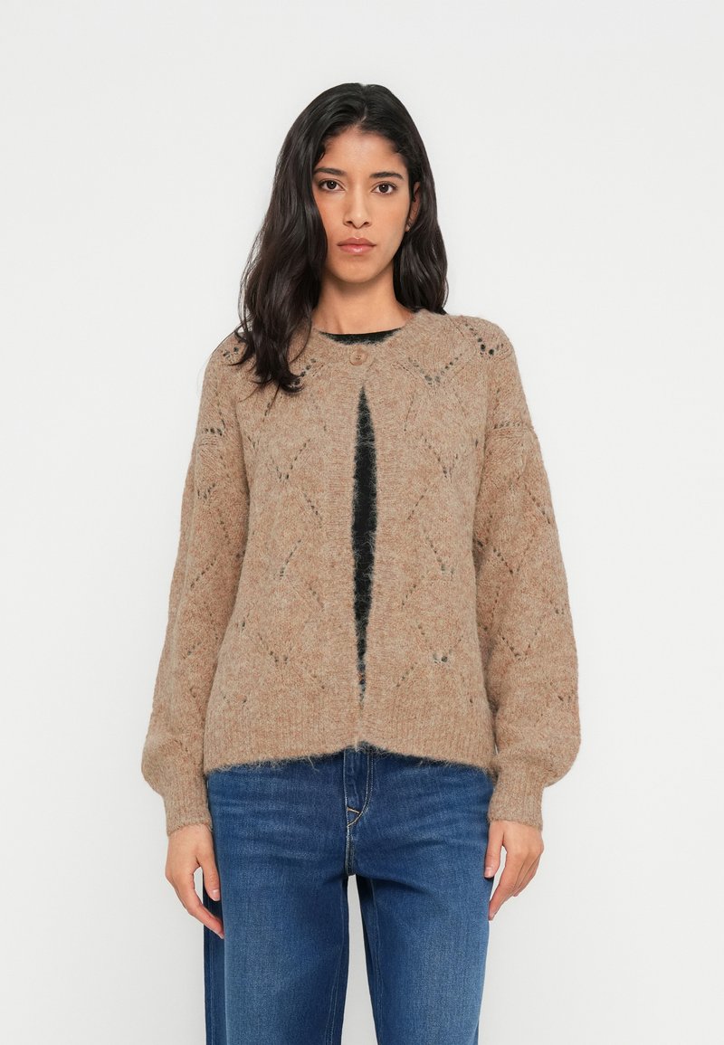 Cardigan beige en tricot avec motif en losange, encolure ronde, fermeture boutonnée et manches volumineuses, porté avec un jean bleu.