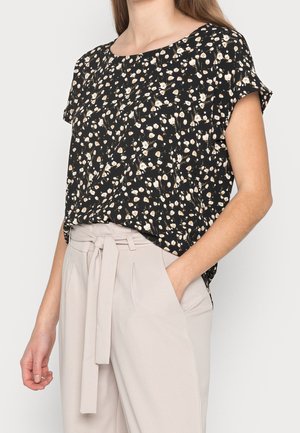 Femme portant un chemisier noir à manches courtes à motif floral, rentré dans un pantalon beige taille haute avec une ceinture en tissu nouée devant.