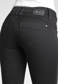 Primer plano de una persona con vaqueros ajustados negros, con bolsillos traseros visibles y una etiqueta de marca en la cintura.