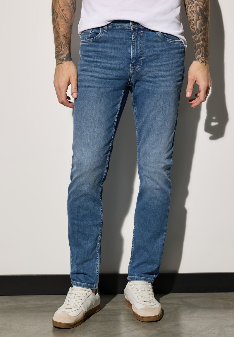 Haalarit vaaleansinisestä farkusta slim fit -mallina, joissa on viisi taskua ja hienovaraisia ompeluyksityiskohtia. Yhdistettynä valkoisiin tennareihin.