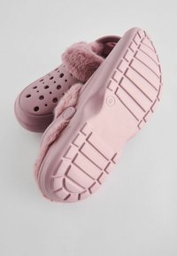 Roze klompen met een bontvoering, voorzien van geperforeerde gaten, een platte zool met een gestructureerd antislippatroon en een gladde afwerking op de bovenkant.