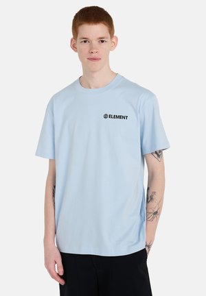 Młoda osoba z rudymi włosami i tatuażami na ramionach ubrana w jasnoniebieską koszulkę z logo Element i czarne spodnie, stojąca na jednolitym białym tle.