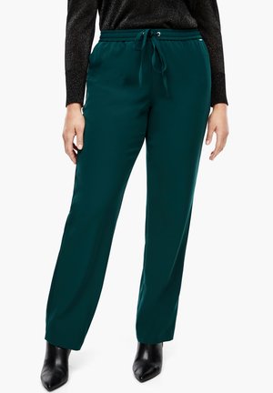Vrouw draagt donkergroene broek met rechte pijpen, een elastische tailleband en een trekkoord, gecombineerd met zwarte enkellaarsjes met een puntige neus.