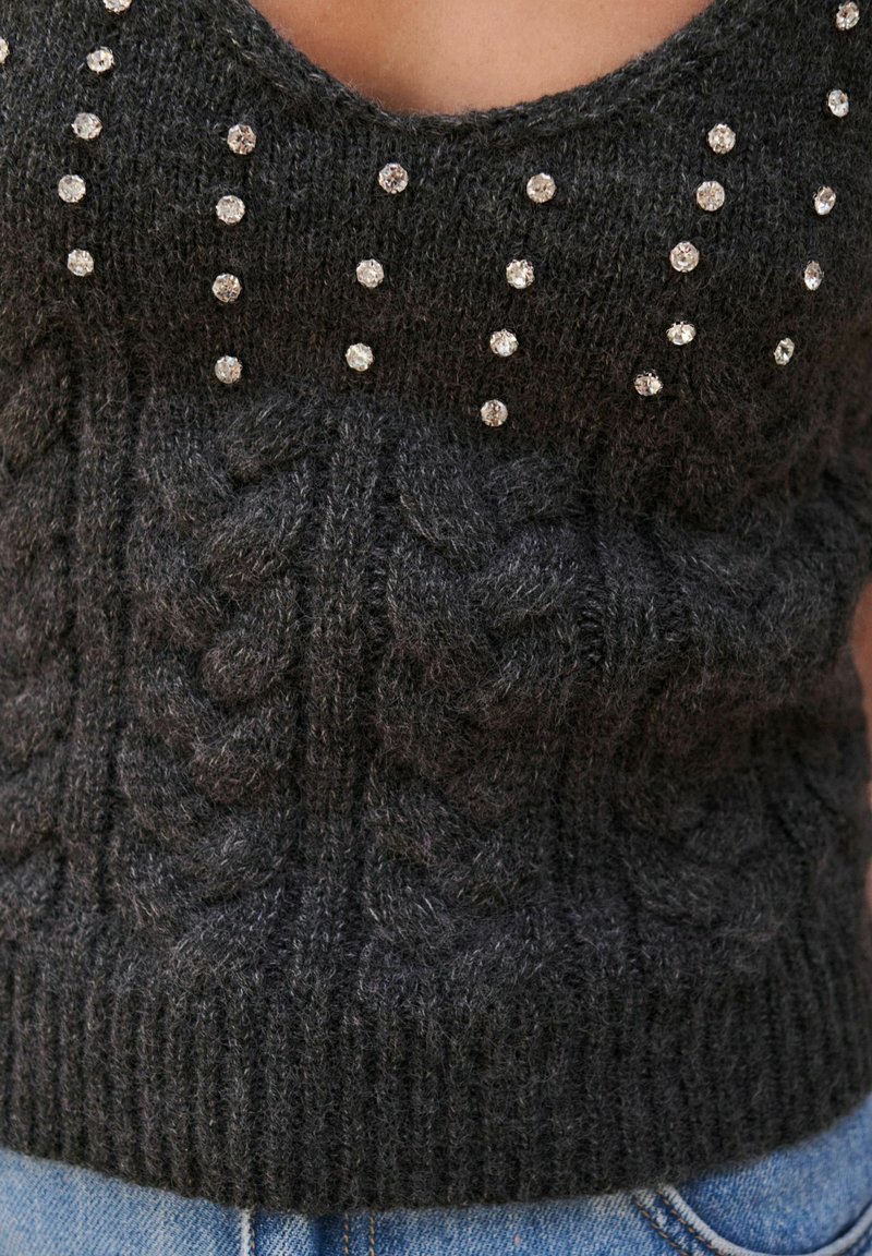 Harmaa neulottu toppi, jossa on syvä V-pääntie, cable-knit -tekstuuri ja jonka kaula-aukkoa koristavat strassit.