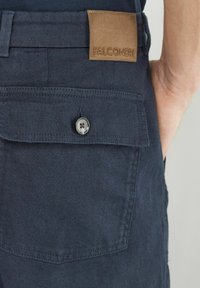 Marinblå byxor i tyg med en läderlapp med texten "FALCONERI", som har en knappstängning på bakfickan och subtila texturdetaljer.