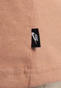 Melna Nike etiķete ar baltu logotipu izšūta persiku krāsā rievojuma audumā tuvu šuvēm un apakšmalai.