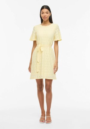 Kobieta o długich ciemnych włosach ubrana w jasną żółtą sukienkę z ażurowym wzorem, krótkimi rękawami, paskiem w talii oraz białe odkryte buty na obcasie, stojąca na białym tle.