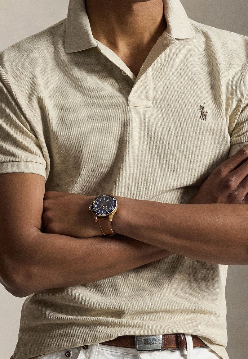 Homme portant un polo beige, les bras croisés, une montre en cuir marron et un pantalon blanc avec une ceinture marron visible.