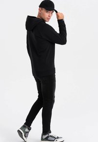Felpa nera con cappuccio e tasca frontale, indossata sopra jeans neri strappati. Un cappello nero è inclinato, e le scarpe sono grigie e bianche con accenti.