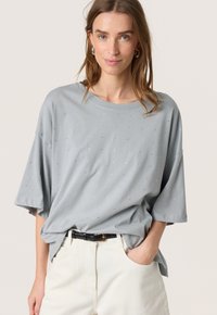 Lys grå oversized t-shirt lavet af blødt stof, med en scoop halsudskæring og tilfældige sølv accenter. Sættes sammen med hvide bukser.