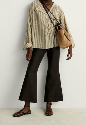 Personne portant un chemisier beige à rayures, un pantalon large noir, des sandales noires, et un sac bandoulière tressé marron clair avec des anses noires.