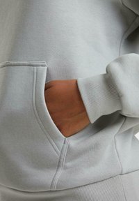 Svetre s kapucňou v svetlosivej farbe, s kloktacou vreckom, rebrovanými manžetami a jemnou textúrou, s viditeľnými detailmi šitia.