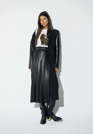 Mujer con falda plisada de cuero negra, abrigo largo negro, camiseta blanca con estampado de leopardo, botas negras con tacones dorados y pendientes de aro dorados.