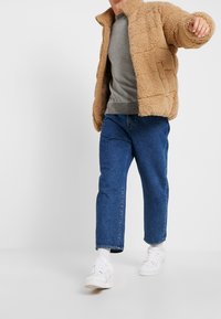 Tan fleece jacket with textured, quilted design, gray sweater underneath, blue straight-leg jeans, and white sneakers. Textured and layered look.