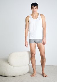 Ein weißes Baumwoll-Trägershirt und graue Boxer-Brief-Unterwäsche, getragen von einem männlichen Modell. Im Hintergrund ist ein weicher, runder, gepolsterter Stuhl in Weiß zu sehen.