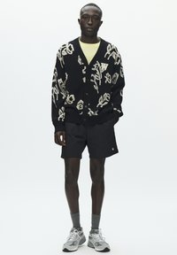 Crni pleteni kardigan s kremastim cvjetnim uzorkom, upareni s crnim kratkim hlačama. Model nosi sive čarape i cipele sa svijetlim detaljima.