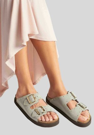 Gambe che indossano sandali beige con due fibbie dorate e borchie, abbinati a un vestito rosa chiaro con orlo asimmetrico.