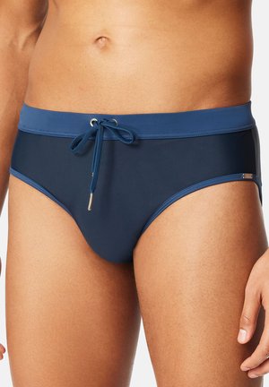 Mies, joka pukeutunut tummansinisiin uimahousuihin, joissa on vaaleansiniset vyötärönauha ja kiristysnauha, seisoo käsi näkyvissä.