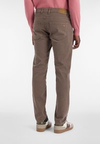 Mann mit braunen karierten Hosen, weißen Sneakers mit grünen und braunen Akzenten und einem rosa Langarmpullover, der vor einem weißen Hintergrund steht.