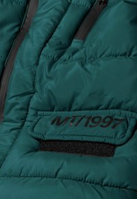 Giacca a vento di colore verde acqua con texture trapuntata. Presenta una patch nera con "MT/1997" e una tasca con zip. Finitura morbida e liscia.
