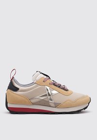 Chaussure de sport beige et tan avec des accents argentés, tige en suède texturé et en tissu, semelle en caoutchouc noir et détail rouge sous le pied.
