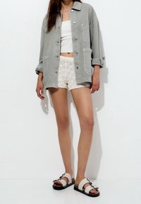 Harmaa nappilukko takki ylipitkällä istuvuudella, valkoinen crop-top, pitsikuosiset shortsit ja kevyet sandaalit, joissa on mustat pohjat ja koristeelliset soljet.