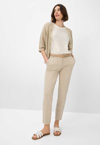 Beige leichter Strickcardigan über einem weißen gestreiften Oberteil, kombiniert mit beigen maßgeschneiderten Hosen und weißen Riemchensandalen.