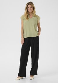 Blusa verde chiaro con maniche corte in pizzo, scollatura a V, abbinata a pantaloni neri a gamba larga e scarpe con tacco cubano beige. Texture liscia, design casual.