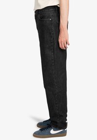 Jean en denim noir présenté dans une coupe droite, montrant des coutures contrastées et des poches avant, associé à des baskets bleues.