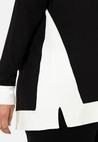 Black and white long-sleeve top with a geometric design, featuring a side slit and contrasting fabric sections. Smooth texture, relaxed fit.