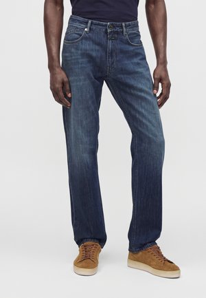 Homme portant un jean droit bleu foncé et des baskets en daim marron, debout sur un fond clair uni.