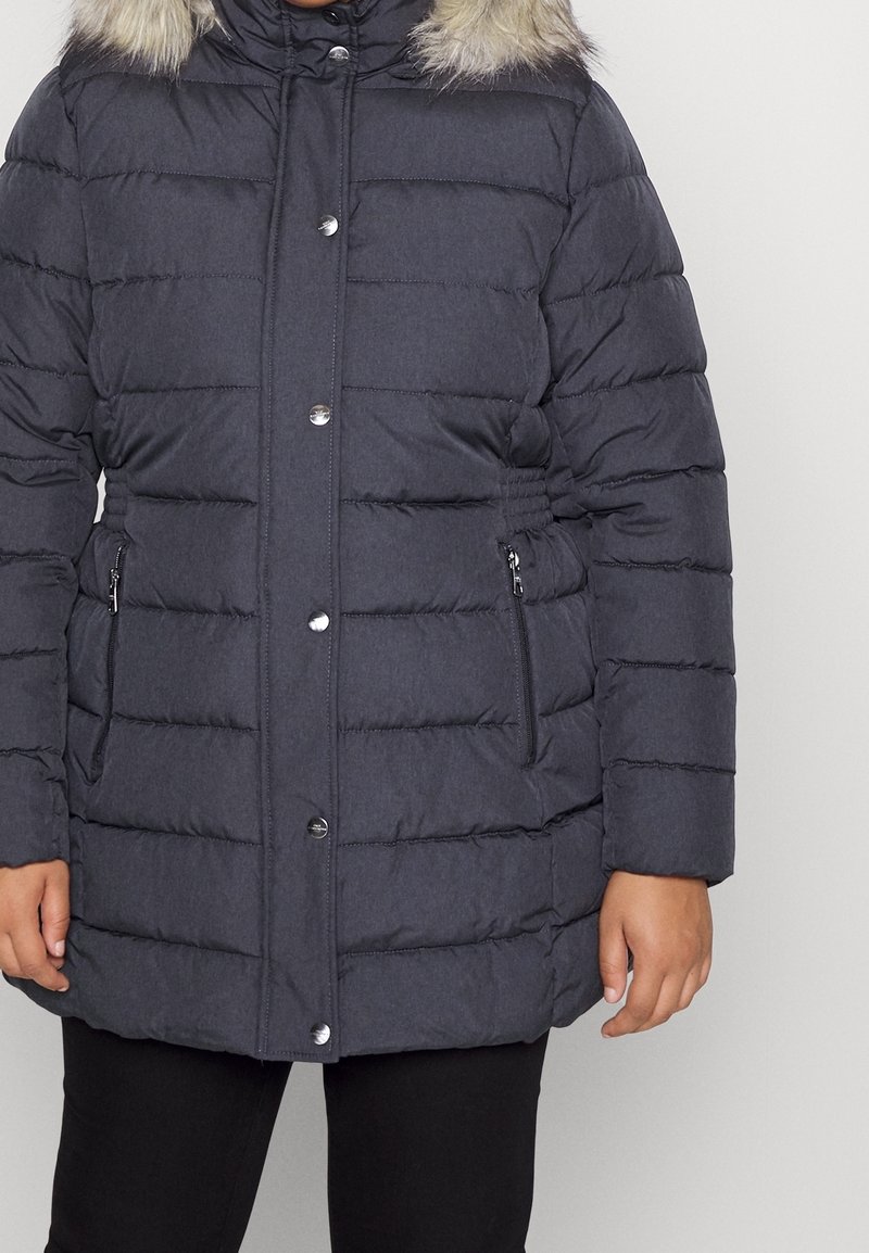 Giacca a vento grigio scuro con sezioni imbottite verticali, cappuccio in finta pelliccia staccabile e due tasche laterali con zip. Bottone a pressione argentato visibile.
