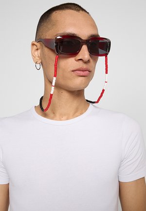 Persona con capelli corti indossa occhiali da sole rettangolari scuri con una catena per occhiali perline rossa, bianca e nera e una maglietta bianca su sfondo grigio.