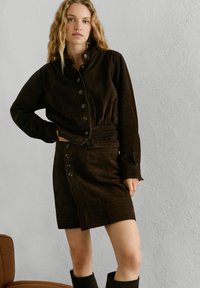 Nuori nainen, jolla on laineikkaat vaaleat hiukset, pukeutuneena tummaan ruskeaan napitettuun takkiin ja siihen sopivaan hameeseen, seisoo vaaleaa, teksturoidun pinnan seinää vasten.