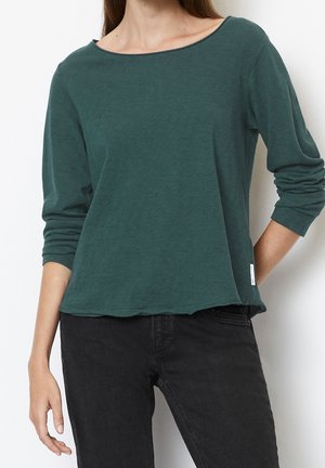 Vrouw draagt een donkergroen langarmige top met bootnek en zwarte jeans, staand met één hand op de heup tegen een witte achtergrond.