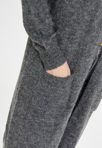 Hand in de zak van een lange, donkergrijze, zachtgebreide cardigan met geribbelde mouwboord.